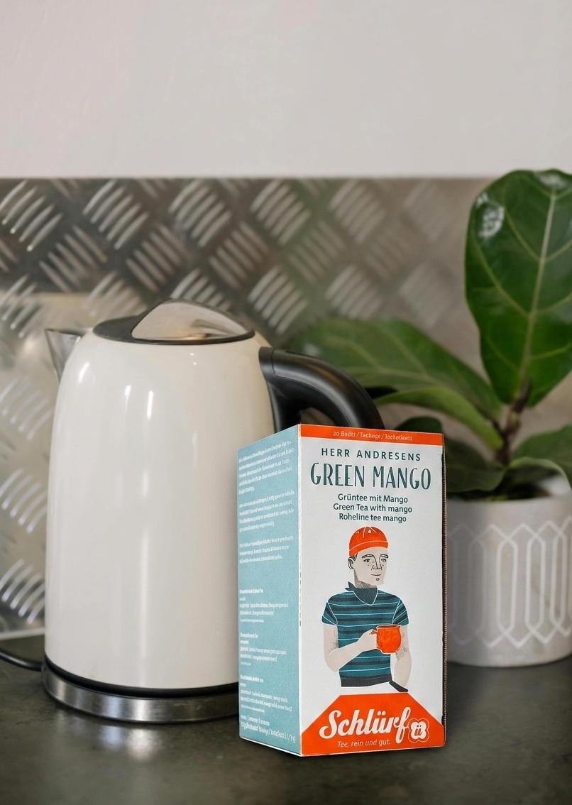 Teeverpackung von dem Schlürf Tee Herr Andresen vor einem Wasserkocher und Blüte
