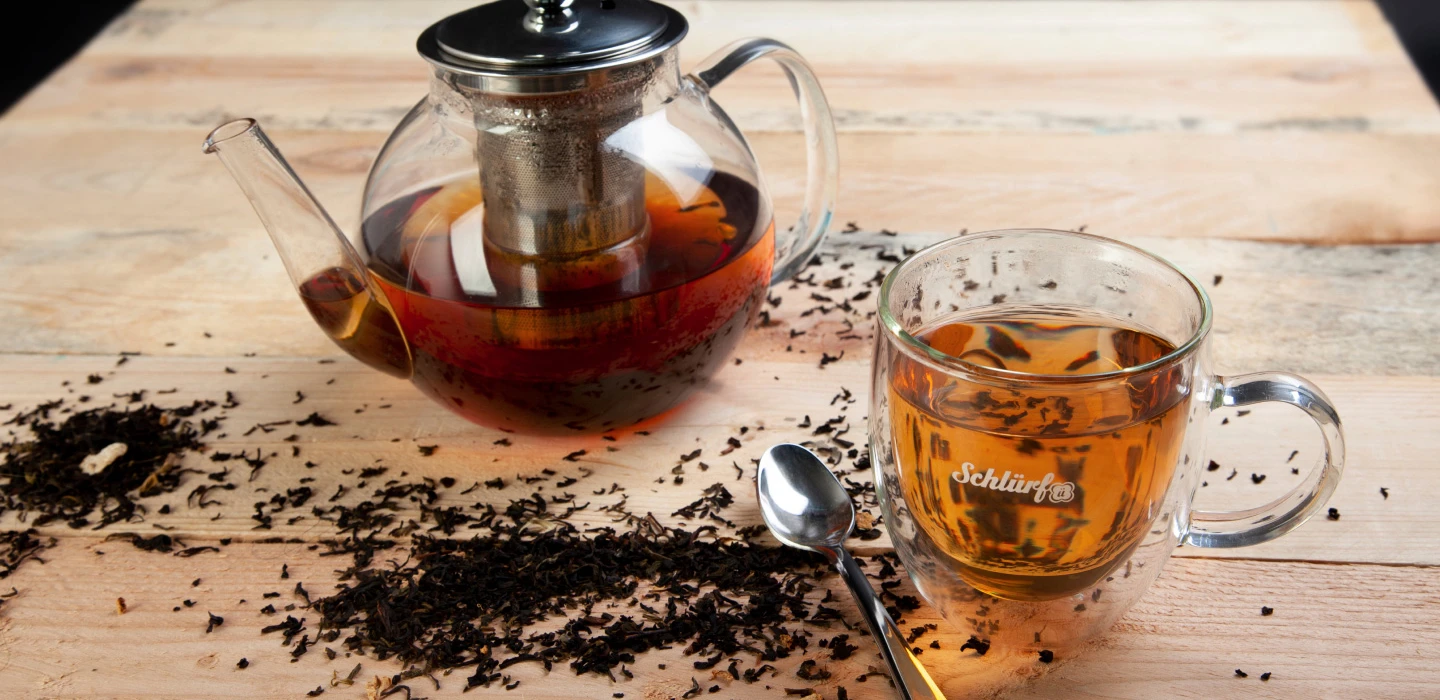 Schlürf Glastasse mit Tee, Teekanne und losem Tee auf Holztisch