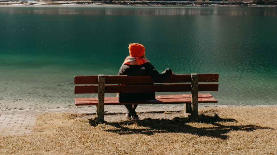 Frau sitzt in der Sonne auf einer Bank und schaut auf einen See